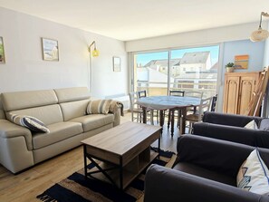 Ein Wohnzimmer mit einer Couch, einem Couchtisch und einem Esstisch mit Stühlen. Ein Fenster bietet Blick auf Gebäude.