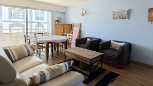 Een woonkamer met een bank, een salontafel, een eettafel met stoelen, een houten kast en een groot raam.