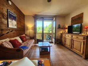 A cozy room with a wooden sofa, a dining table, a TV, and a balcony with a view of greenery. - Quiet apartment, pets allowed - 4 guests (Samoëns)