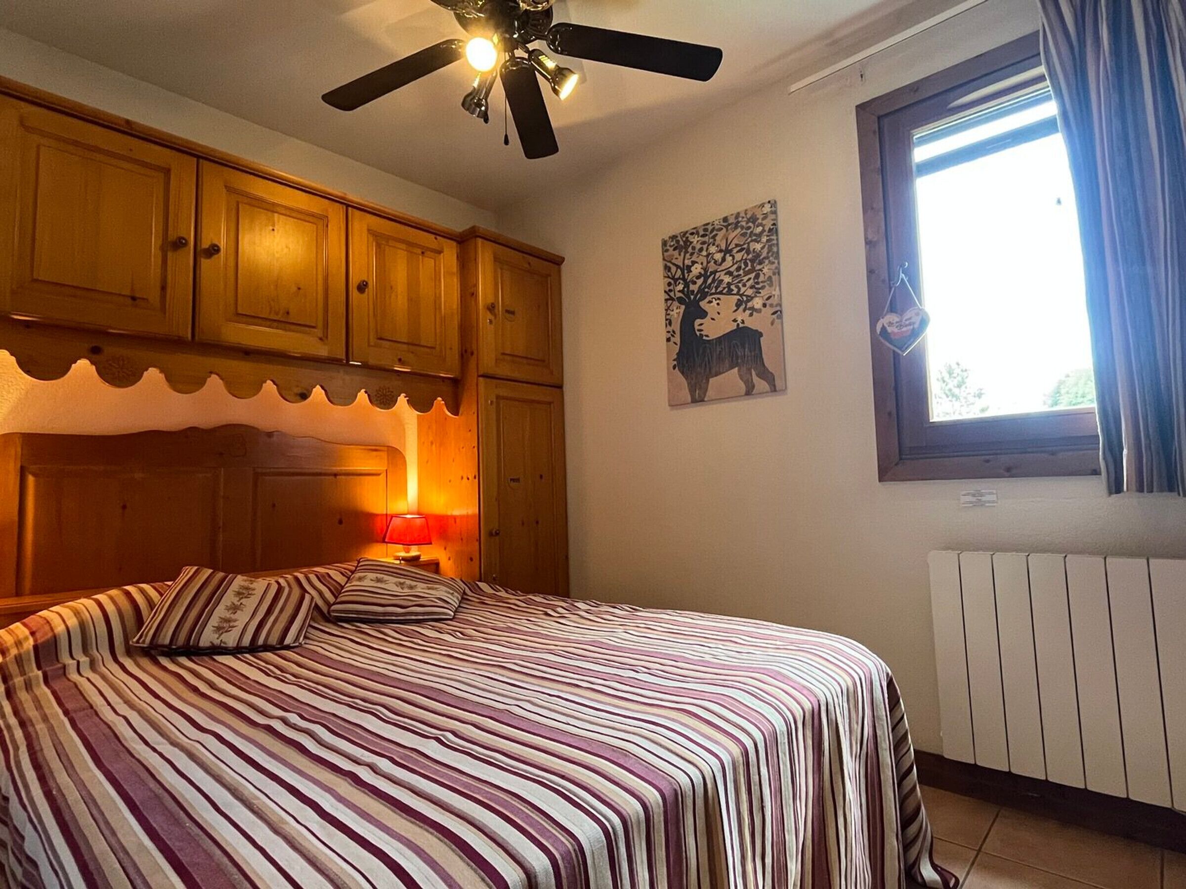 Ein Schlafzimmer mit einem Bett, Holzschränken, einem Deckenventilator, einem Fenster mit Vorhängen und einem an der Wand befestigten Heizkörper.