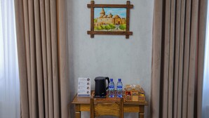 A room with a painting of a building, a table with a kettle, water bottles, and a sign. - Shergiron Plaza (Bukhara)