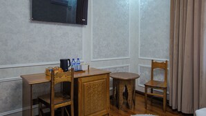 A hotel room with a flat-screen TV mounted on the wall, a wooden table with chairs, a small round table, and a bottle of water. - Shergiron Plaza (Bukhara)