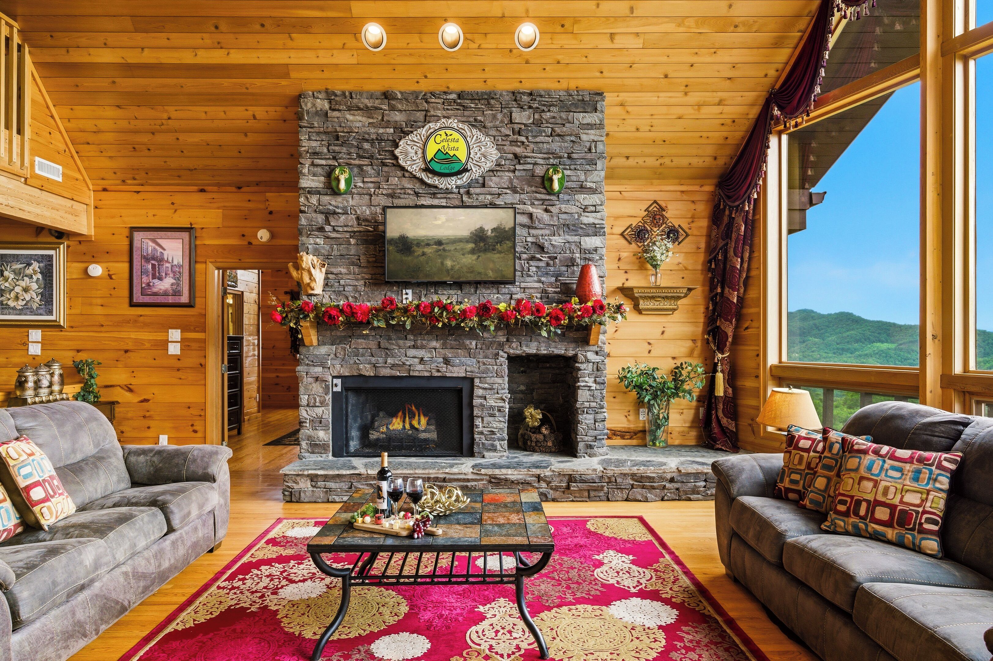 A cozy living room with a stone fireplace, wooden walls, and a large window offering a scenic view.