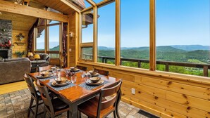 Ein Holzinterieur mit einem für vier Personen gedeckten Esstisch, großen Fenstern mit Blick auf die Berge und einem Kamin.