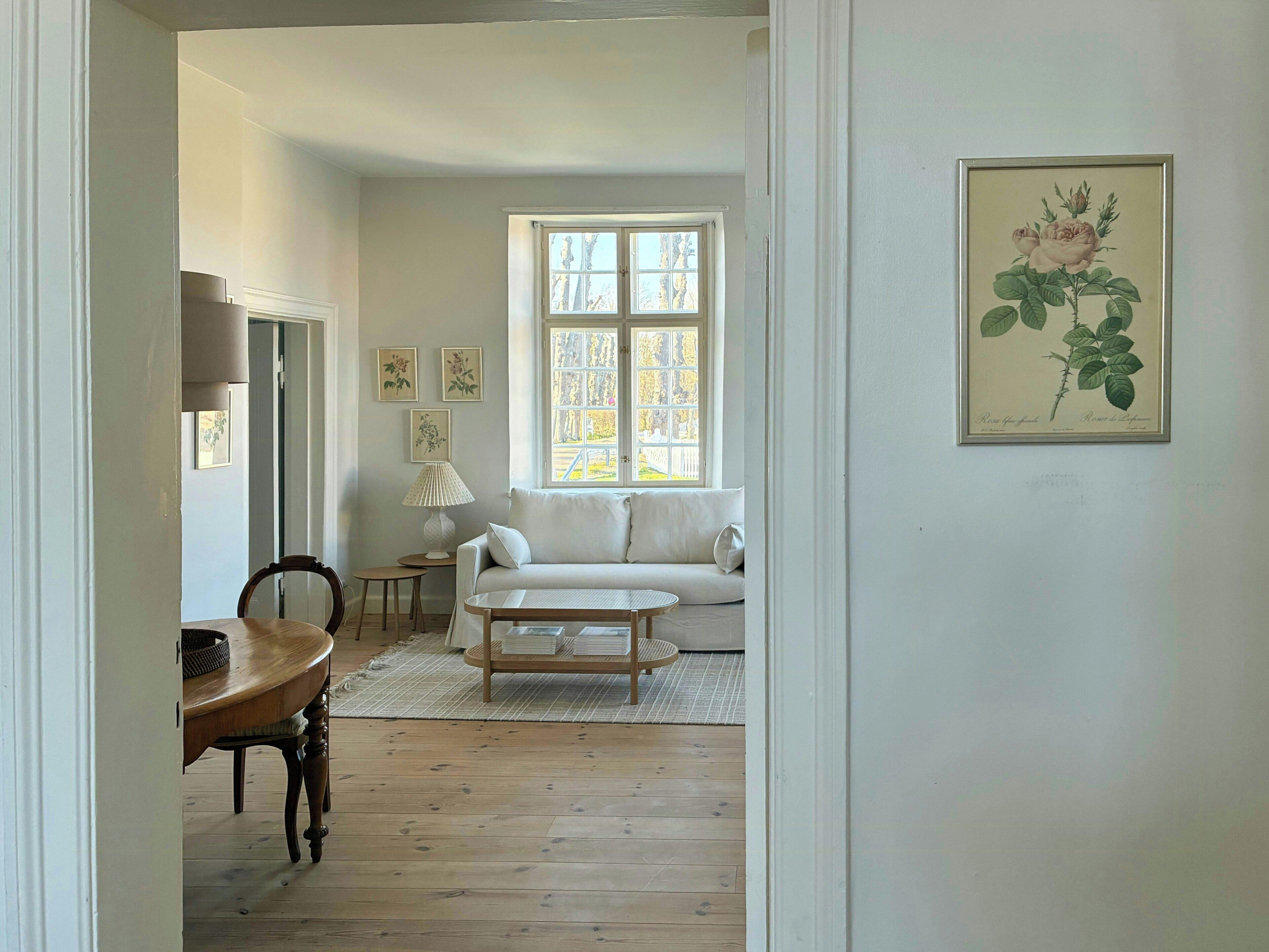 Ein Zimmer mit Holzboden, einem weißen Sofa, einem kleinen runden Tisch und einem Fenster mit Blick auf Bäume.