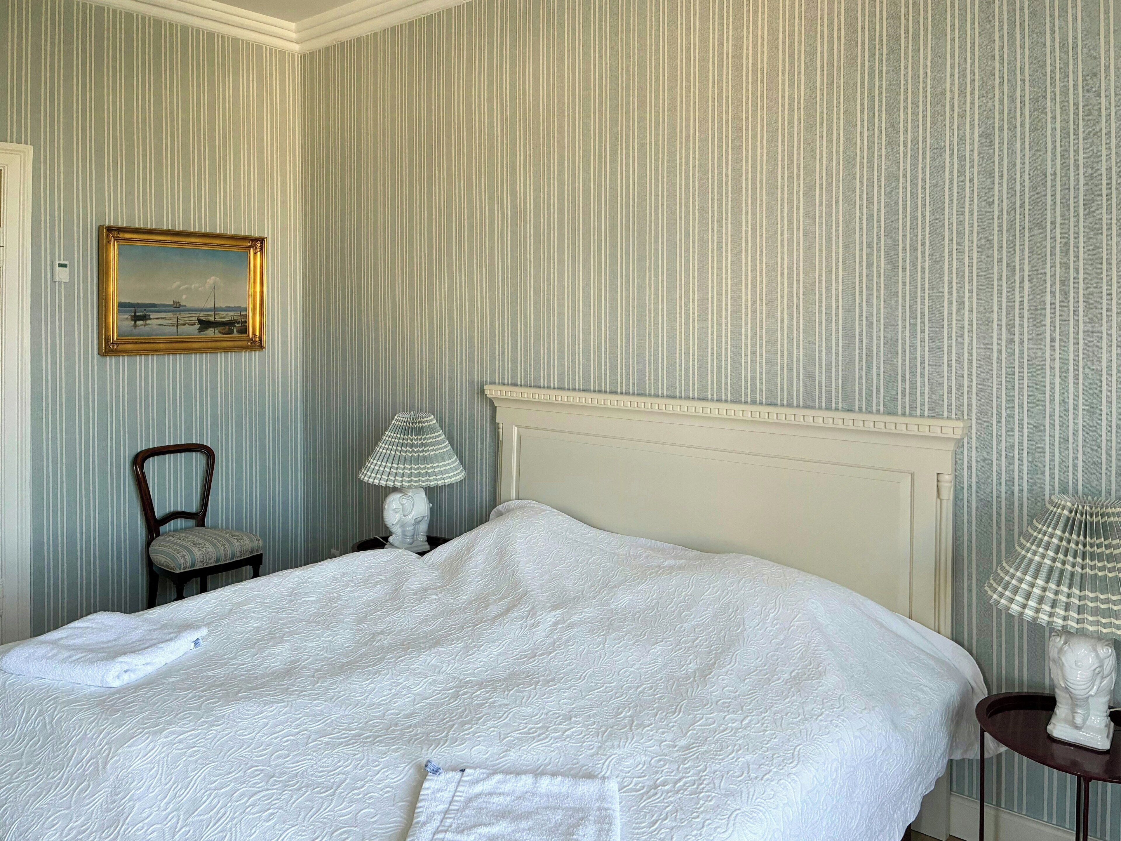 Un lit bien fait, recouvert d’une couette blanche, une chaise, deux lampes et un tableau encadré au mur.