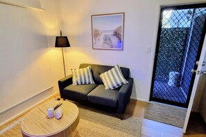 Una sala de estar con un sofá, dos almohadas, una lámpara, una mesa redonda con tazas y un cuadro enmarcado en la pared.