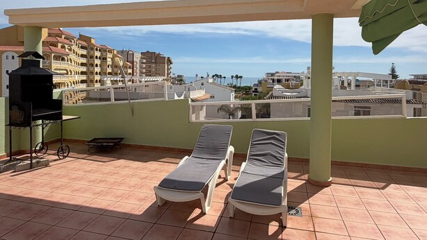 Terraço no telhado com duas espreguiçadeiras, churrasqueira e vista para edifícios e o mar.