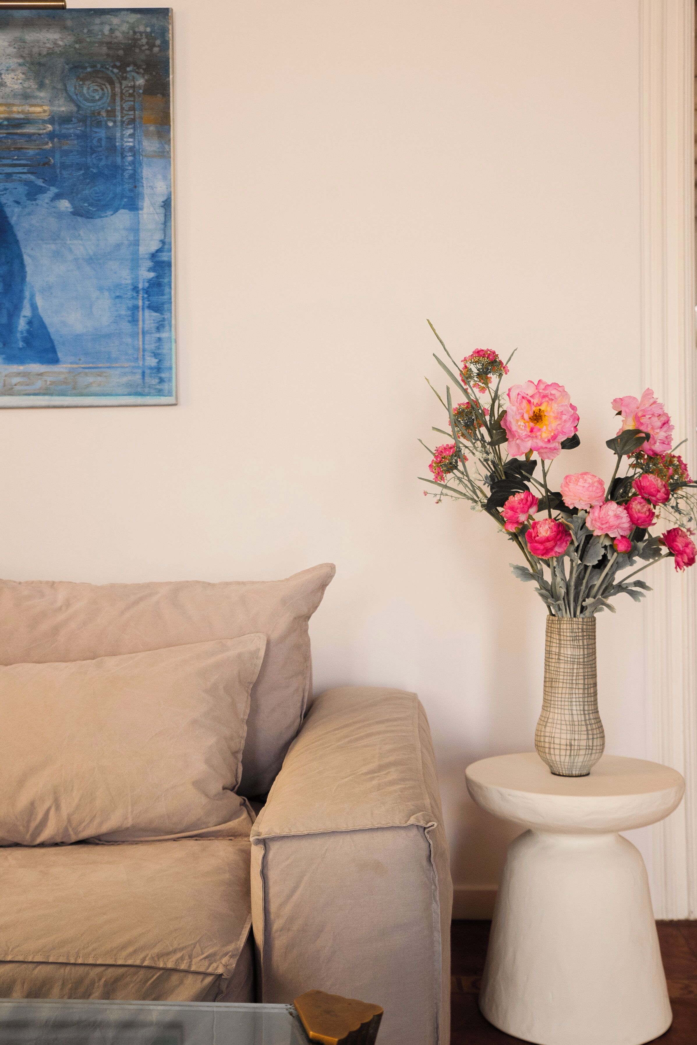 A beige sofa, a white side table, and a vase with pink flowers.