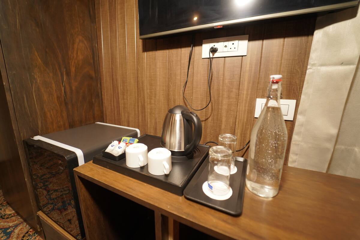 Quarto de hotel com parede de madeira, televisão de tela plana fixada acima de uma tomada, uma mesinha com chaleira, duas canecas brancas, dois copos e uma garrafa de água.