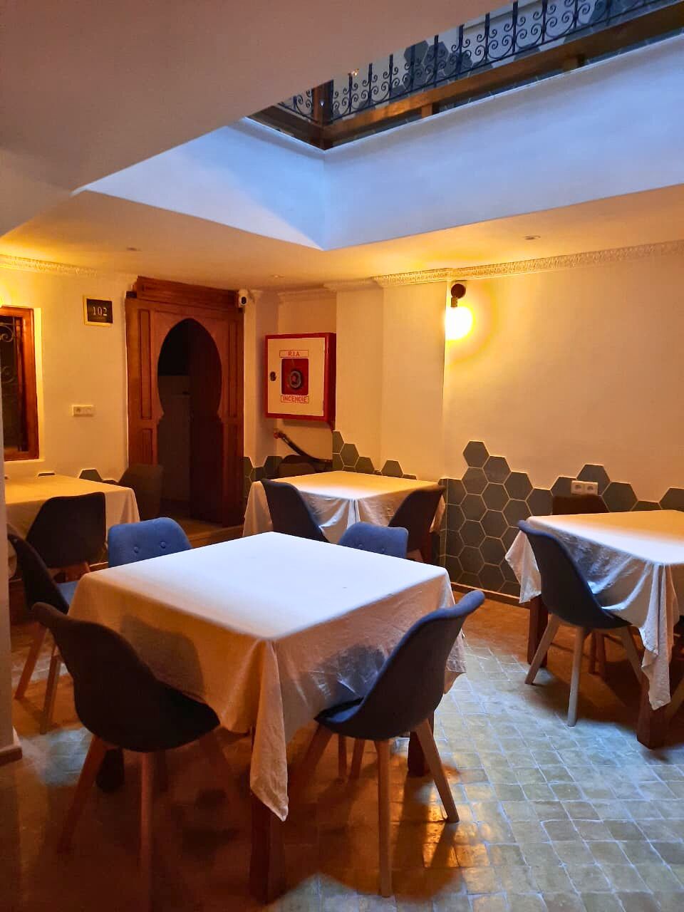 Un comedor con mesas y sillas, un techo con tragaluces y una lámpara fijada a la pared.