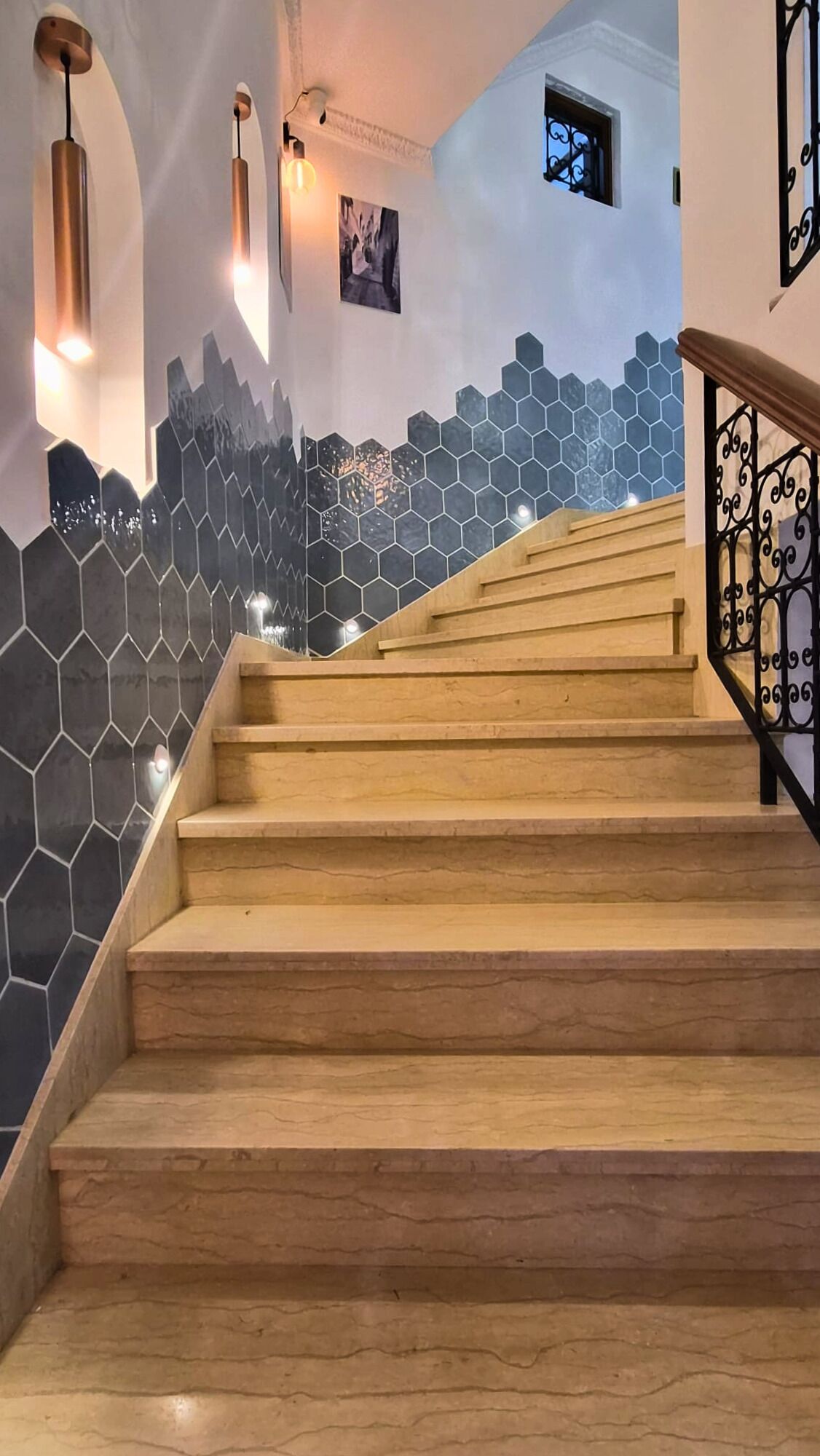 Un escalier en bois, avec un mur recouvert de carreaux hexagonaux noirs et blancs, une petite fenêtre et une rambarde en métal.