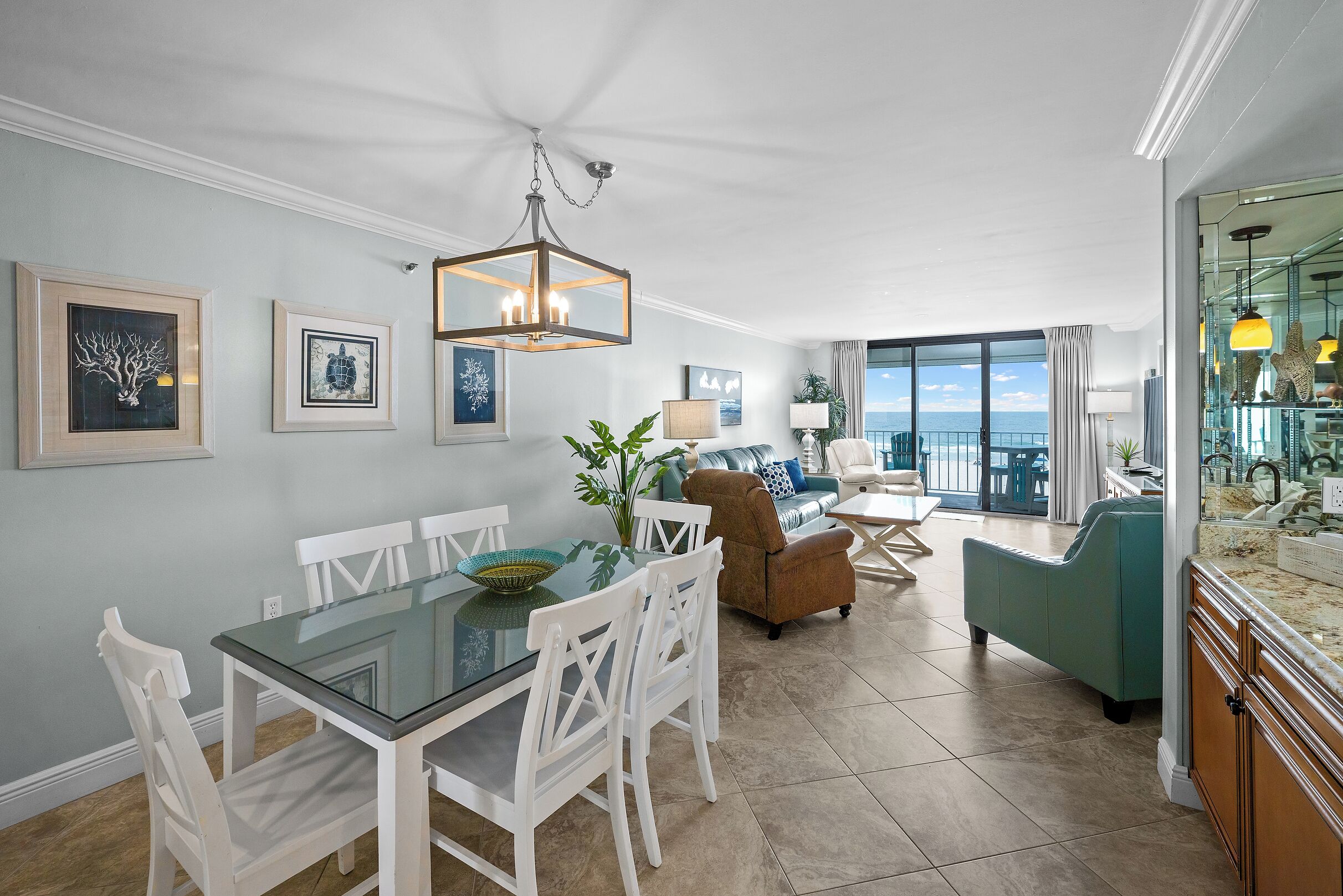 A dining area with a glass table and white chairs, a chandelier, framed artwork on the wall, and a view of the ocean through large windows.