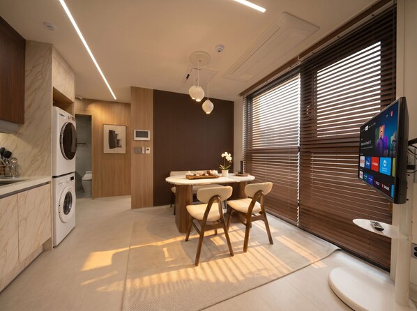 Une cuisine moderne équipée d’un lave-linge intégré, un coin repas avec une table ronde et des chaises, et un grand téléviseur fixé au mur.