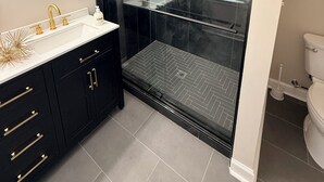 A modern bathroom with a glass-enclosed shower, a round mirror, a gold faucet, and black cabinetry. - Welcome to Sandtown Villa—your home away from home. (Atlanta)