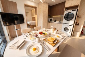 A modern kitchen with a dining area, a flat-screen TV, a microwave, a washing machine, and a breakfast setup with eggs, toast, and fruit. - PRANA I  Full-Service APT in Gangnam 5 min. Metro (SEOUL)