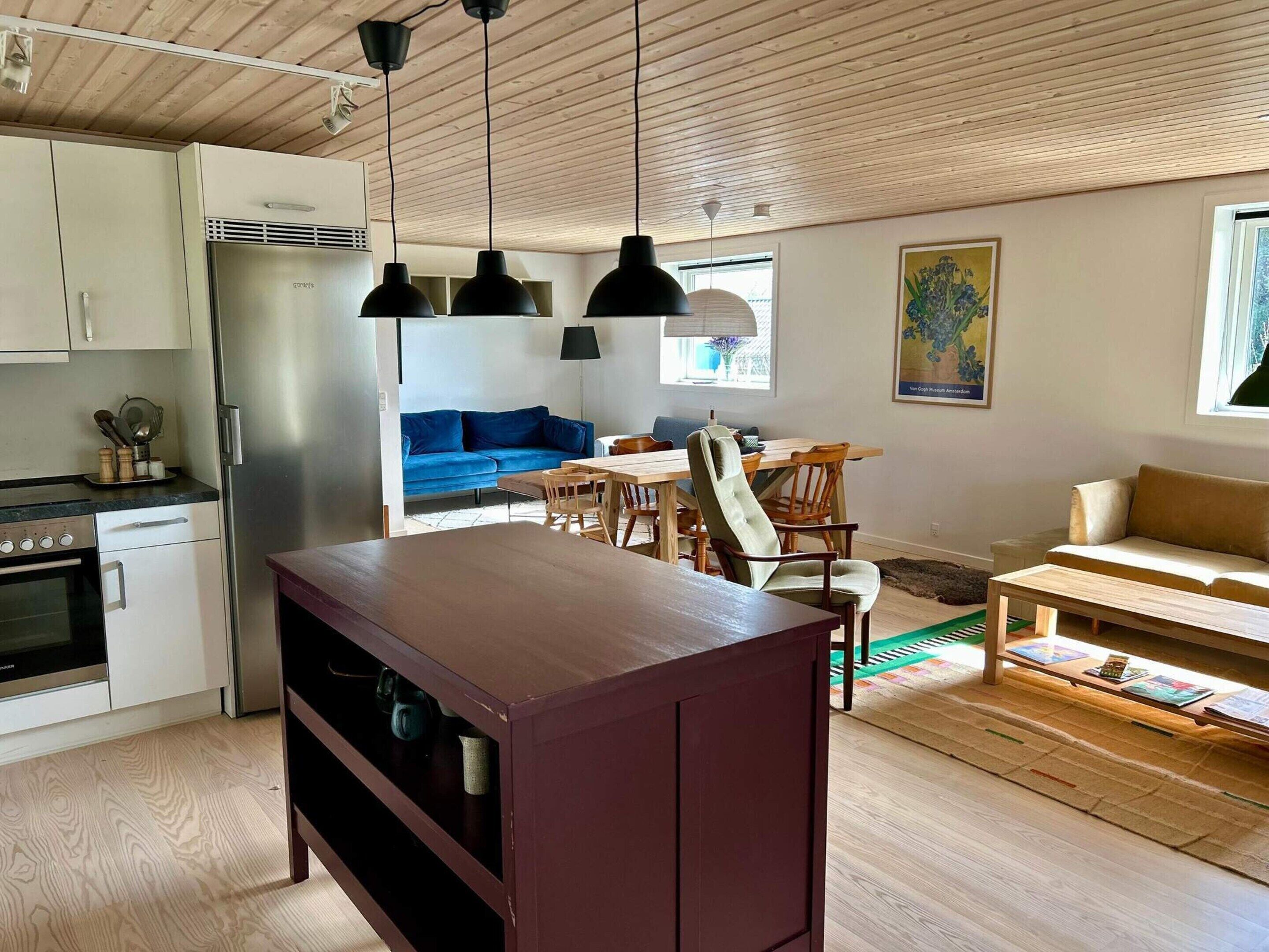 Uma cozinha moderna com forno embutido e geladeira, uma sala de jantar com móveis de madeira e uma sala de estar com sofá azul e sofá bege.