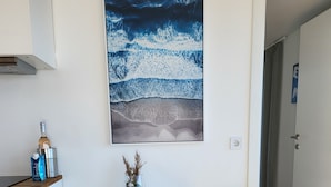 Ein weißer Schreibtisch mit zwei gelben Stühlen, einem gerahmten Kunstwerk mit einer Darstellung einer Welle und einer kleinen Vase mit Trockenpflanzen.