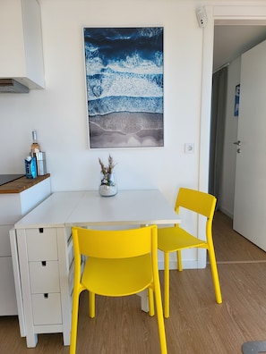 A white desk with two yellow chairs, a framed artwork of a wave, and a small vase with dried plants.