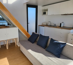A compact living space with a kitchenette, a sofa with cushions, a wooden staircase, and a view through a door.