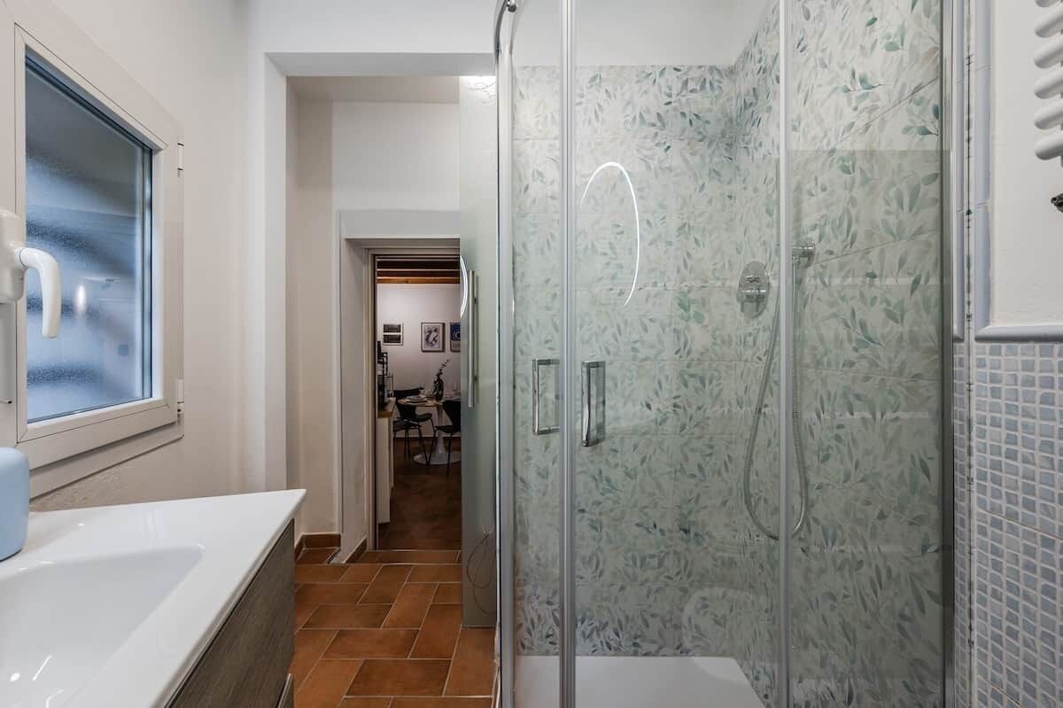 A modern bathroom with a glass shower enclosure, a white sink, and a visible hallway leading to a dining area.