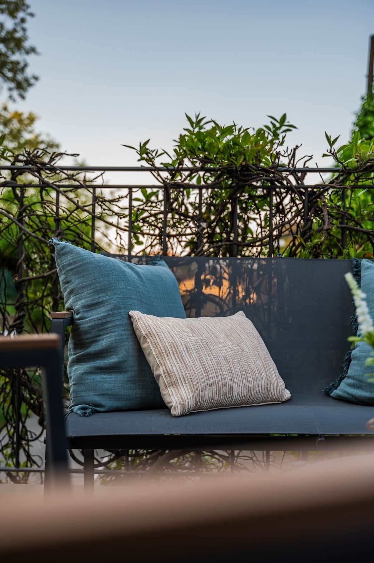 Un canapé d’extérieur noir, agrémenté de deux coussins, un bleu et un beige, placé devant une clôture en métal, avec une végétation luxuriante en arrière-plan.
