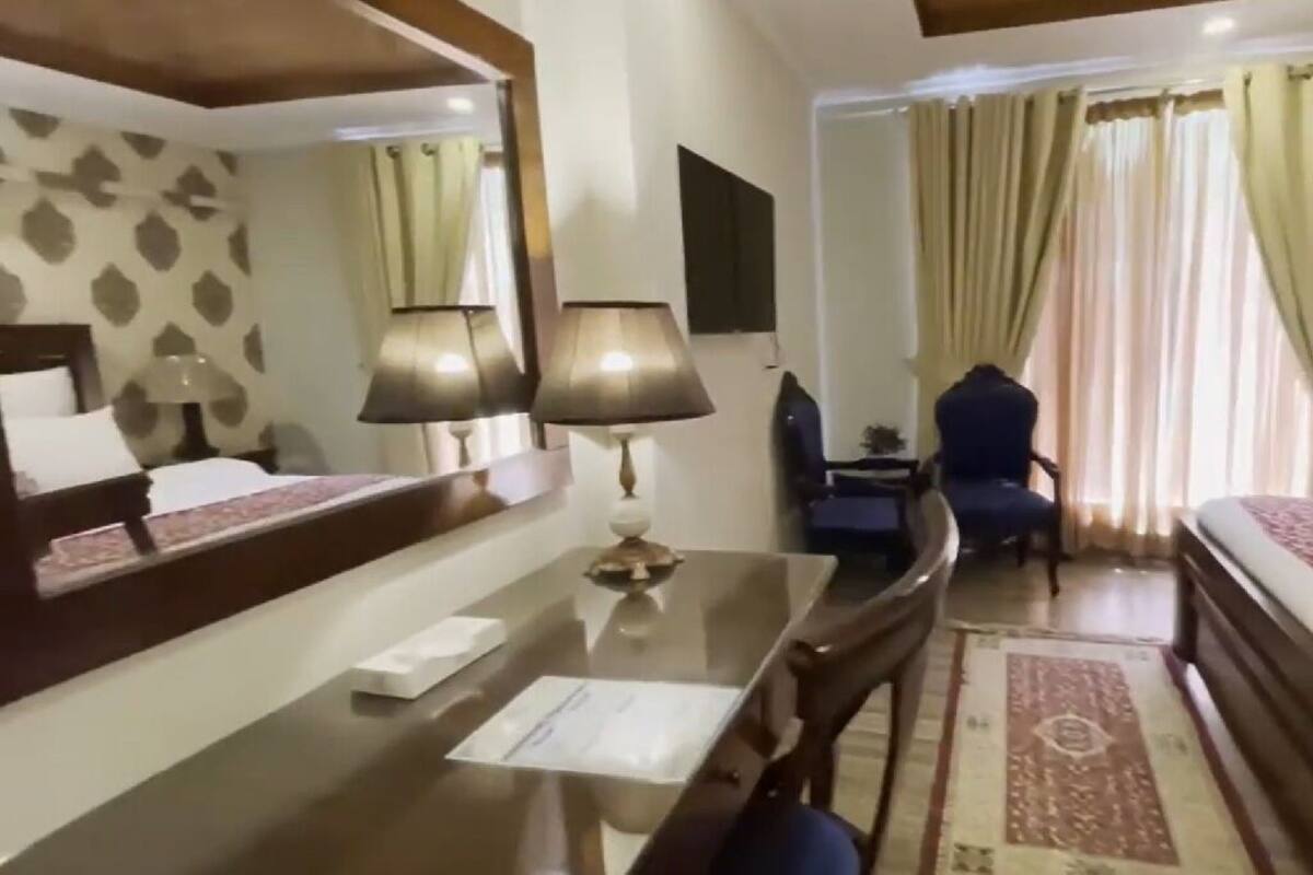 A hotel room with a bed, a desk with a lamp, a chair, a small table, and a mirror.