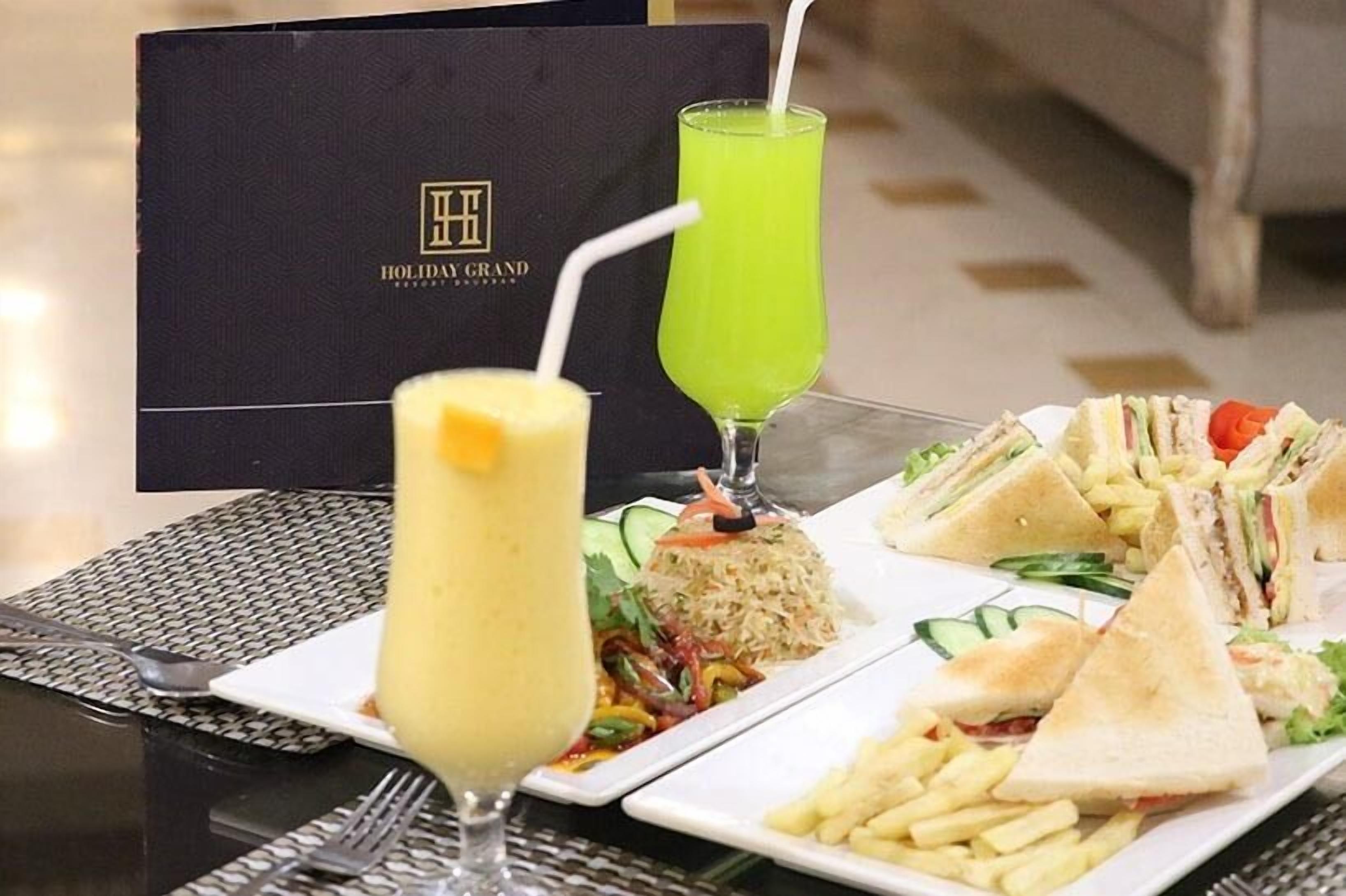 Ein Tisch mit einem Getränk, Sandwiches und Pommes, sowie eine schwarze Box mit dem Text 'Holiday Grand'.
