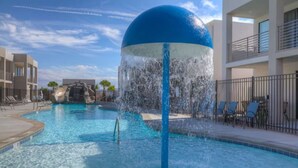 A swimming pool with a water fountain feature, lounge chairs, and a building with balconies in the background. - The Lofts #17 Townhome Pool Resort, Poolside and Hot tub (St. George)