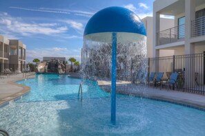 A swimming pool with a water fountain feature, lounge chairs, and a building with balconies in the background. - The Lofts #17 Townhome Pool Resort, Poolside and Hot tub (St. George)