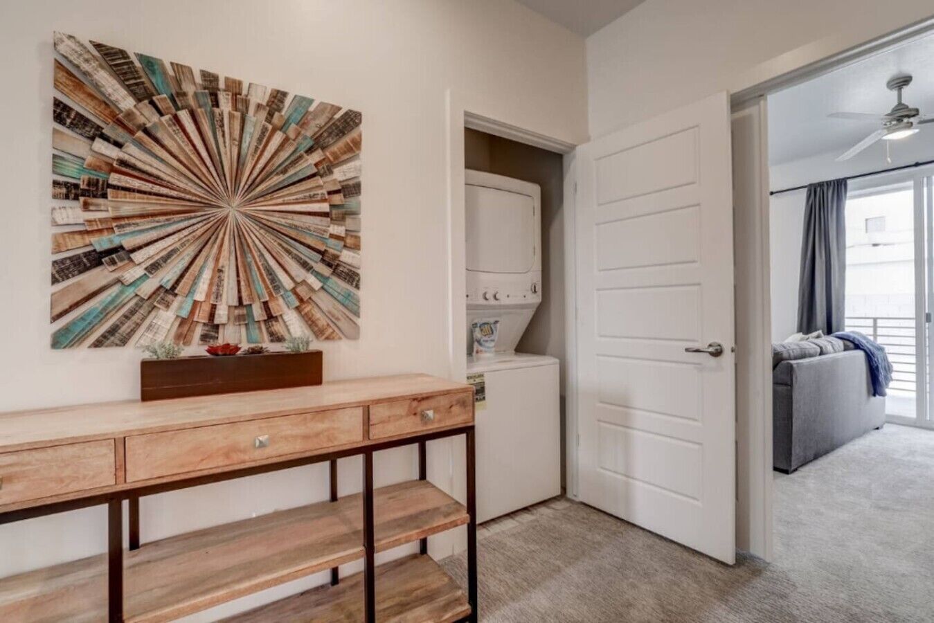 A room with a wooden console table, a wall-mounted abstract art piece, a white door, and a grey sofa in the background.