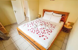 A neatly made bed with rose petals and a towel arrangement, a wooden headboard, a bedside lamp, and a door in the background. - La Villa Siana - A getaway between sea and nature (Sainte-Suzanne)