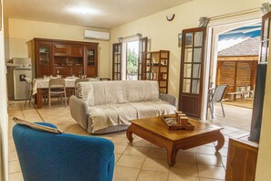 Ein Wohnzimmer mit einer Couch, einem Couchtisch und einem blauen Sessel. Es gibt einen Essbereich mit Tisch und Stühlen sowie eine Küchenzeile mit Schränken und einem Kühlschrank.