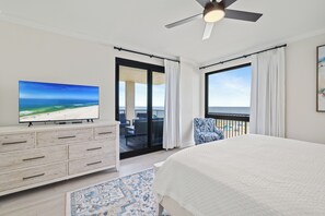 A bedroom with a bed, a TV, a dresser, a balcony with a view of the beach, and a ceiling fan.