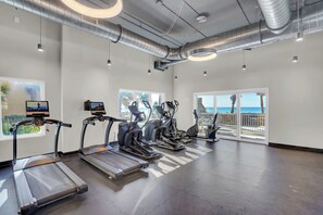 Ein Fitnessraum mit Laufbändern, Crosstrainern und einem Rudergerät. Große Fenster bieten Blick auf Palmen und den Ozean.