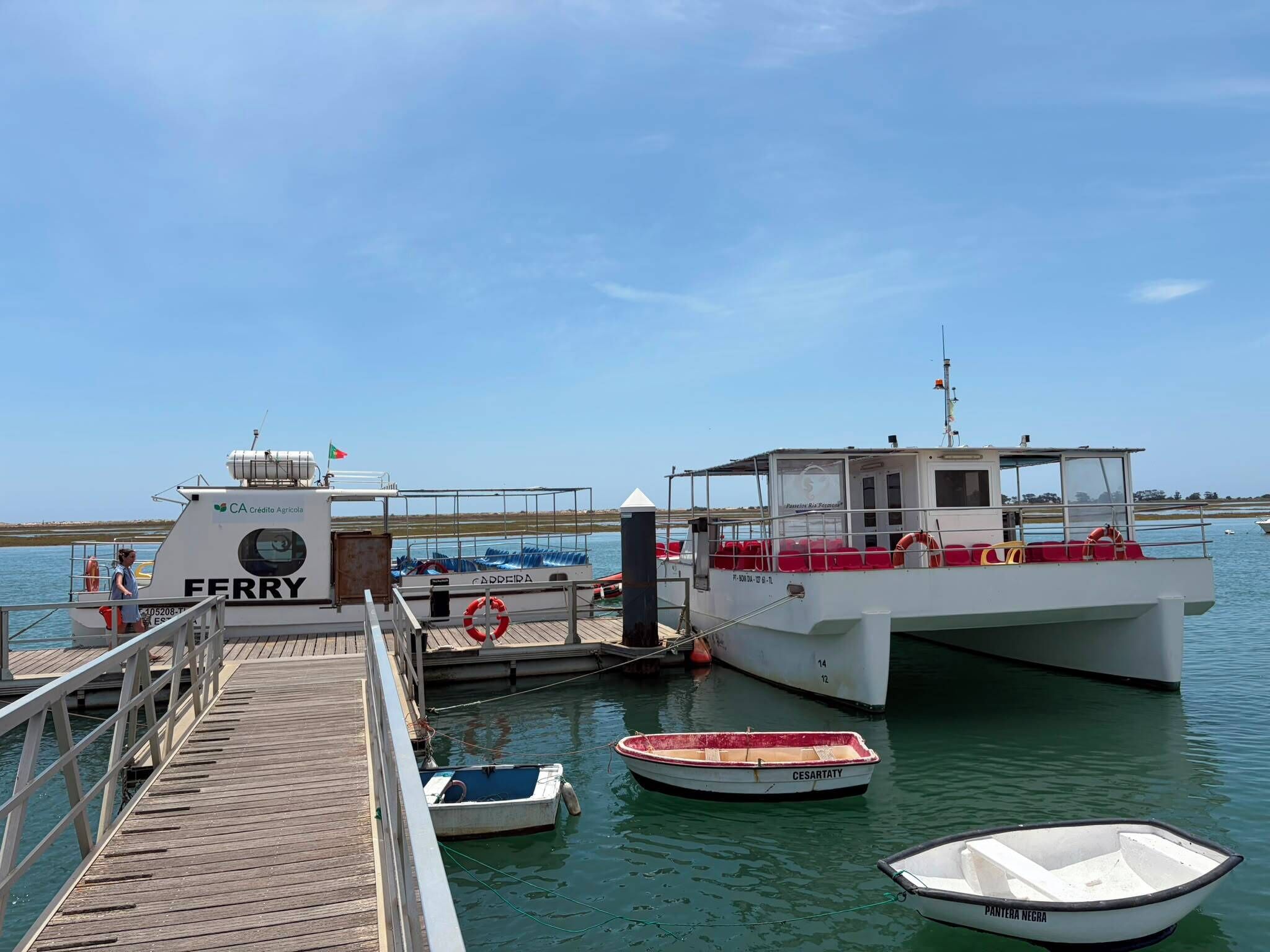 A ferry dock with a wooden pier, a white ferry named 'FERRY,' and two small boats named 'CESARTATY' and 'PANTERA NEGRA.'