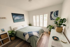 Ein Schlafzimmer mit einem Bett, einem Fenster mit Jalousien, einem Gemälde und mehreren Blumentöpfen.