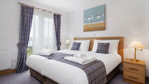 Une chambre d’hôtel avec un lit, une table de chevet avec une lampe, un tableau représentant une scène de plage et une fenêtre avec des rideaux.