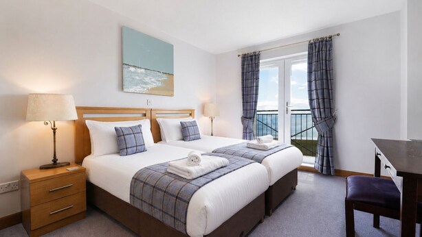 Une chambre d’hôtel avec deux lits, un bureau, une chaise, une table de chevet, une lampe et une vue sur la mer par la fenêtre.