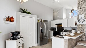 A modern kitchen with white cabinetry, a stainless steel refrigerator, a microwave, and a stone accent wall. - Lake Serenity Retreat Private Pool, Lake Views, Game Room & Outdoor TV (Kissimmee)