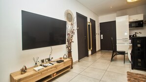 A modern living room with a flat-screen TV, a wooden console table, a vase with flowers, a mirror, and a dining area with a chair and table. - Cozy 1-bedroom apartment in SIGNATURE (Accra)