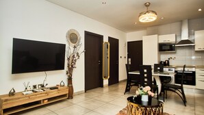 A modern living room with a flat-screen TV, a wooden entertainment unit, a dining area with a table and chairs, and a kitchen with white cabinets and a microwave. - Cozy 1-bedroom apartment in SIGNATURE (Accra)