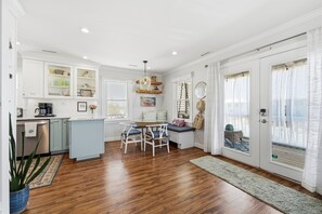 A modern kitchen with white cabinets, a dining area with a round table and chairs, and a large window with curtains. - Deja Blue - North Topsail Beach Rental with Tiki Bar & Outdoor Living (North Topsail beach)