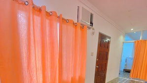 Ein Zimmer mit Holzboden, orangefarbenen Vorhängen, einem Bett mit gestreifter Bettdecke und einer Tür zu einem weiteren Raum.