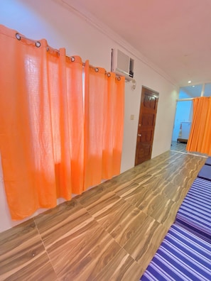 A room with a wooden floor, orange curtains, a bed with a striped bedspread, and a door leading to another room.