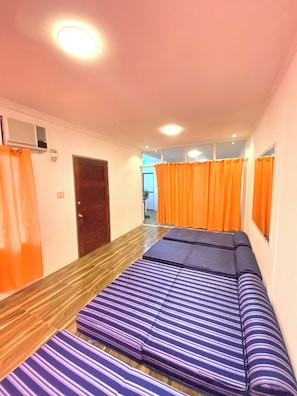 A room with a wooden floor, a bed with striped bedding, orange curtains, and a ceiling light.