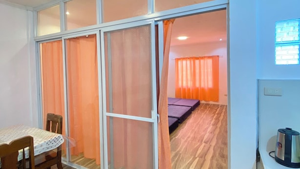 Ein Zimmer mit Esstisch, Stühlen, einem Bett mit orangefarbenen Vorhängen, einer Whiteboard-Tafel und einem Wasserkocher auf einer Arbeitsplatte.