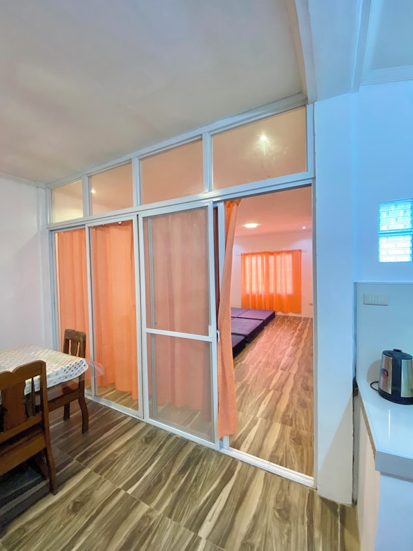 A room with a dining table, chairs, a bed with orange curtains, a whiteboard, and a kettle on a counter.