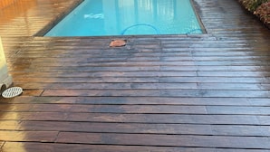 Ein Schwimmbad mit Holzterrasse, ein Haus mit rotem Ziegeldach und ein kleiner Garten.