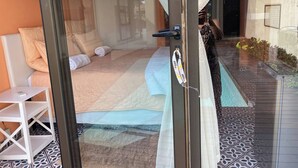 Ein Zimmer mit Bett, kleinem Tisch, Stuhl, Glastür und Blick ins Freie.